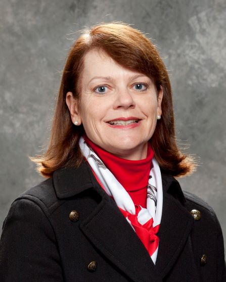 Professional portrait of a woman wearing a black coat and red scarf, smiling against a textured gray background. Suitable for a business profile.